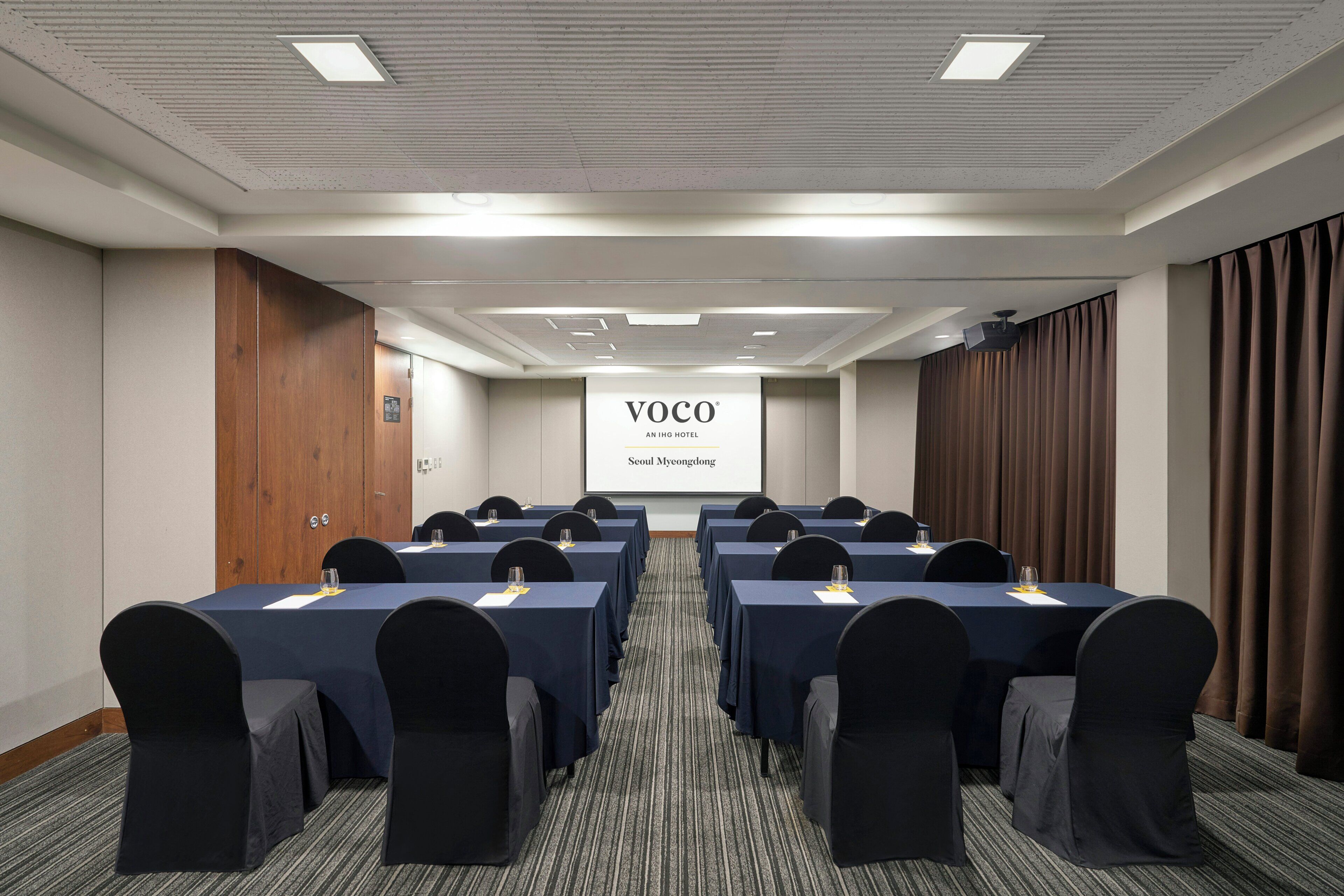 Un salón de conferencias con varias mesas y sillas, una pantalla de proyección mostrando 'VOCO AN IHG HOTEL Seoul Myongdong' y una puerta de madera a la izquierda.
