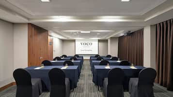 Un salón de conferencias con varias mesas y sillas, una pantalla de proyección mostrando 'VOCO AN IHG HOTEL Seoul Myongdong' y una puerta de madera a la izquierda.