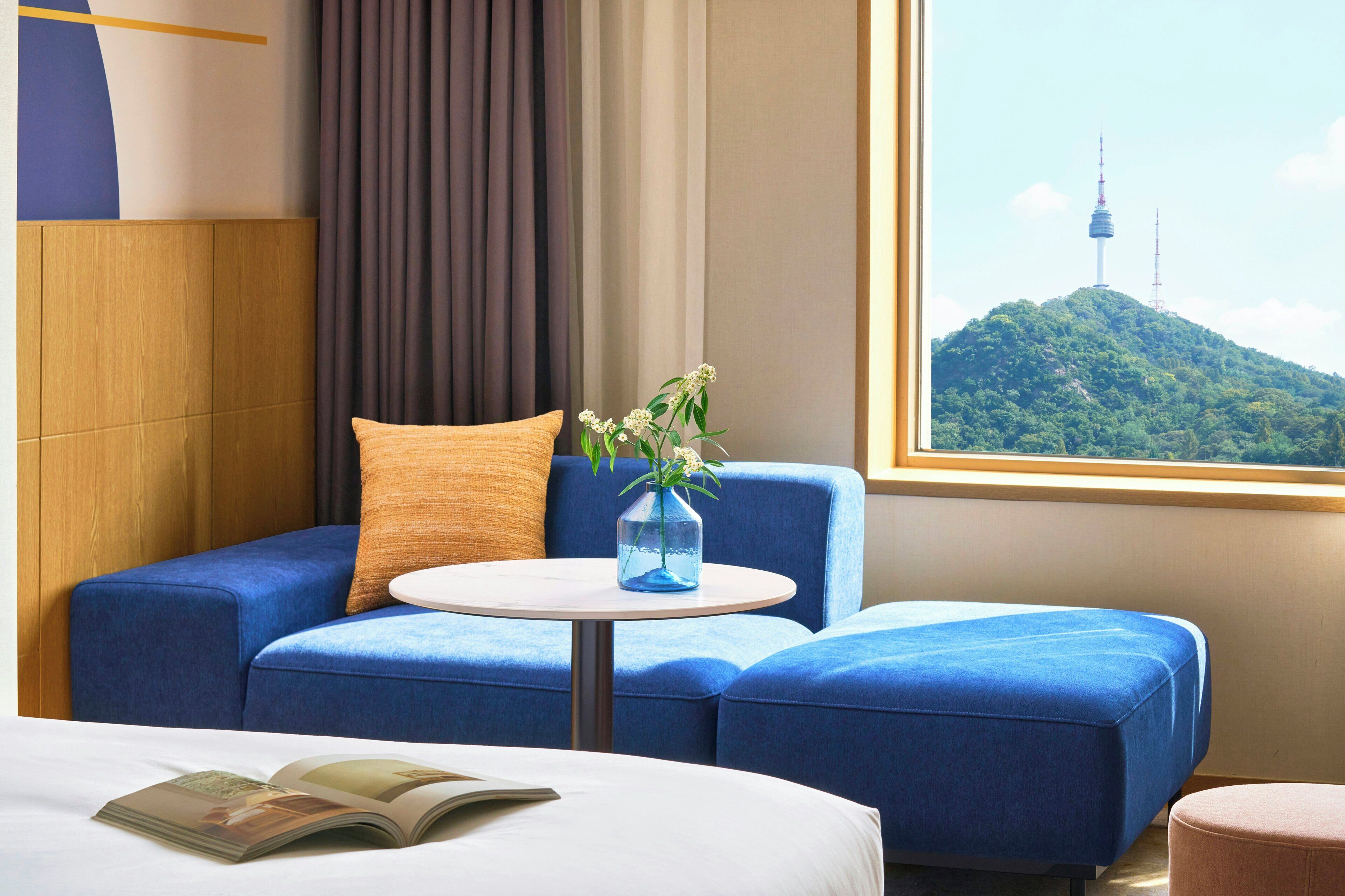 A hotel room with a blue sofa, a small round table, and a view of a mountain with a tower.
