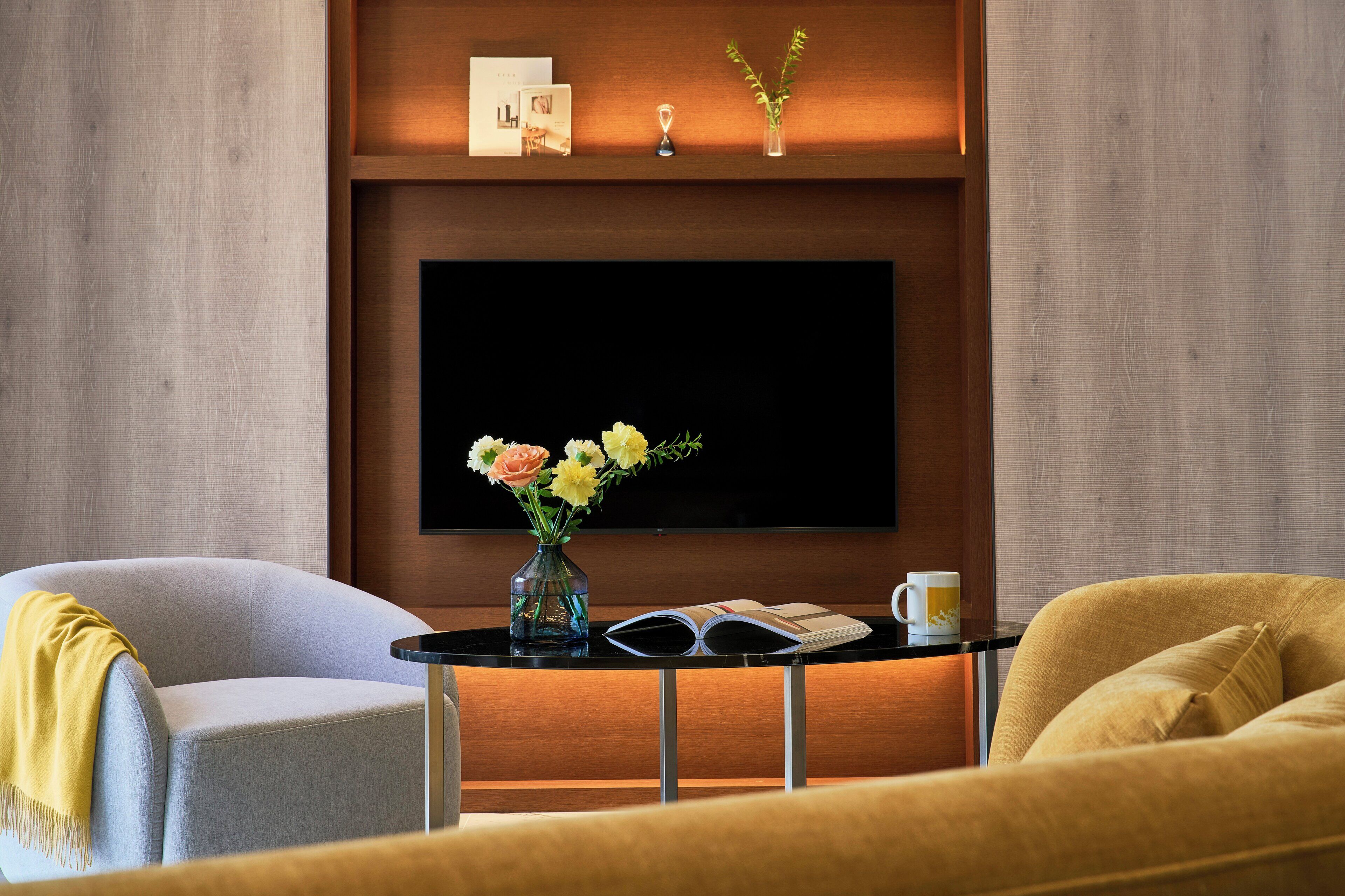 A modern living room with a flat-screen TV mounted on a wooden wall, a small table with a vase of flowers, and two armchairs.