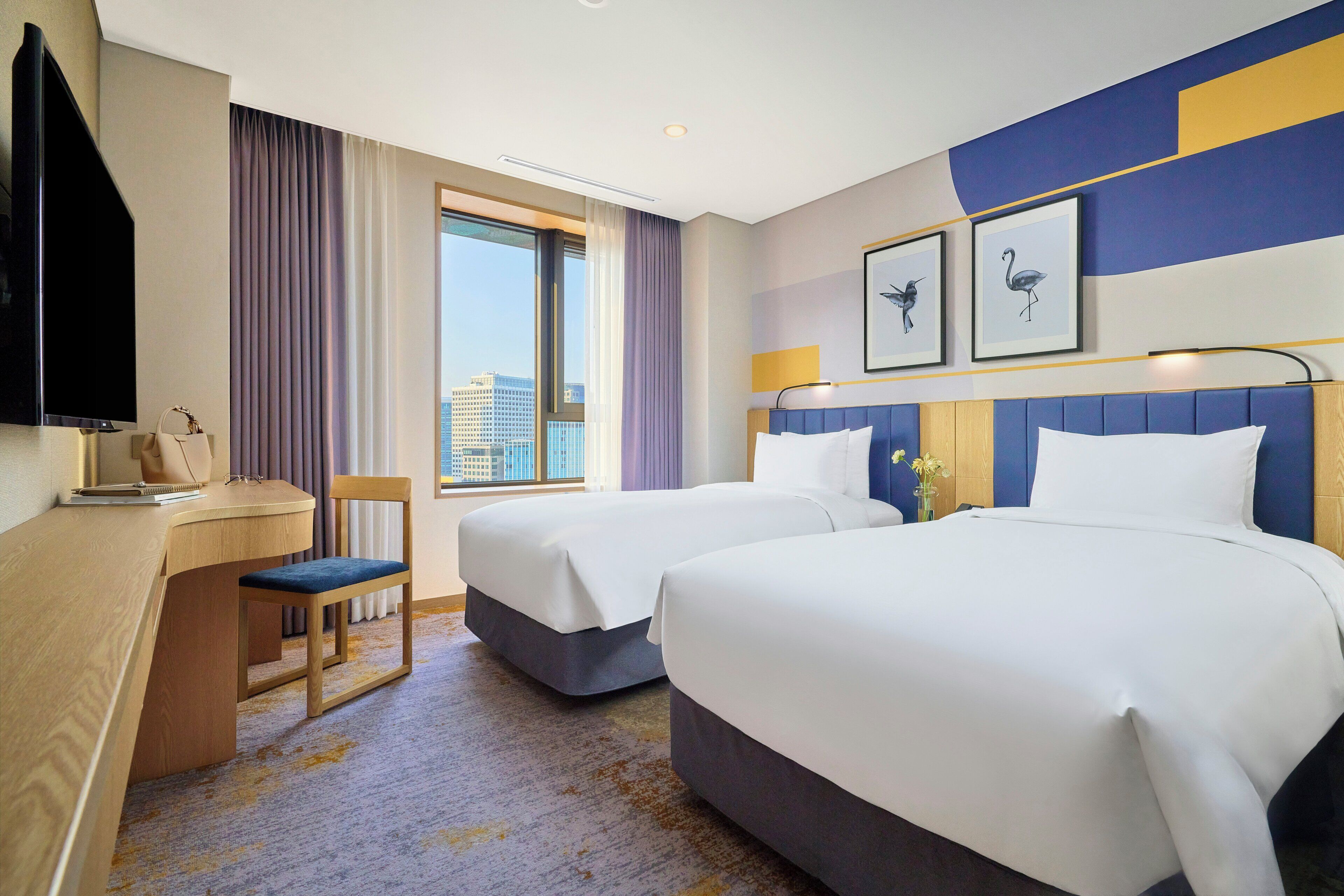 A hotel room with two beds, a desk, a chair, a television, and a window with a city view.