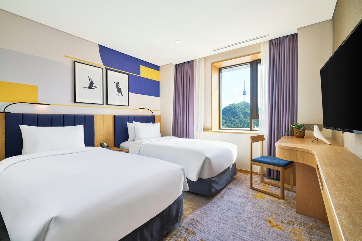 A hotel room with two beds, a desk, a TV, and a window with a view of a tower.