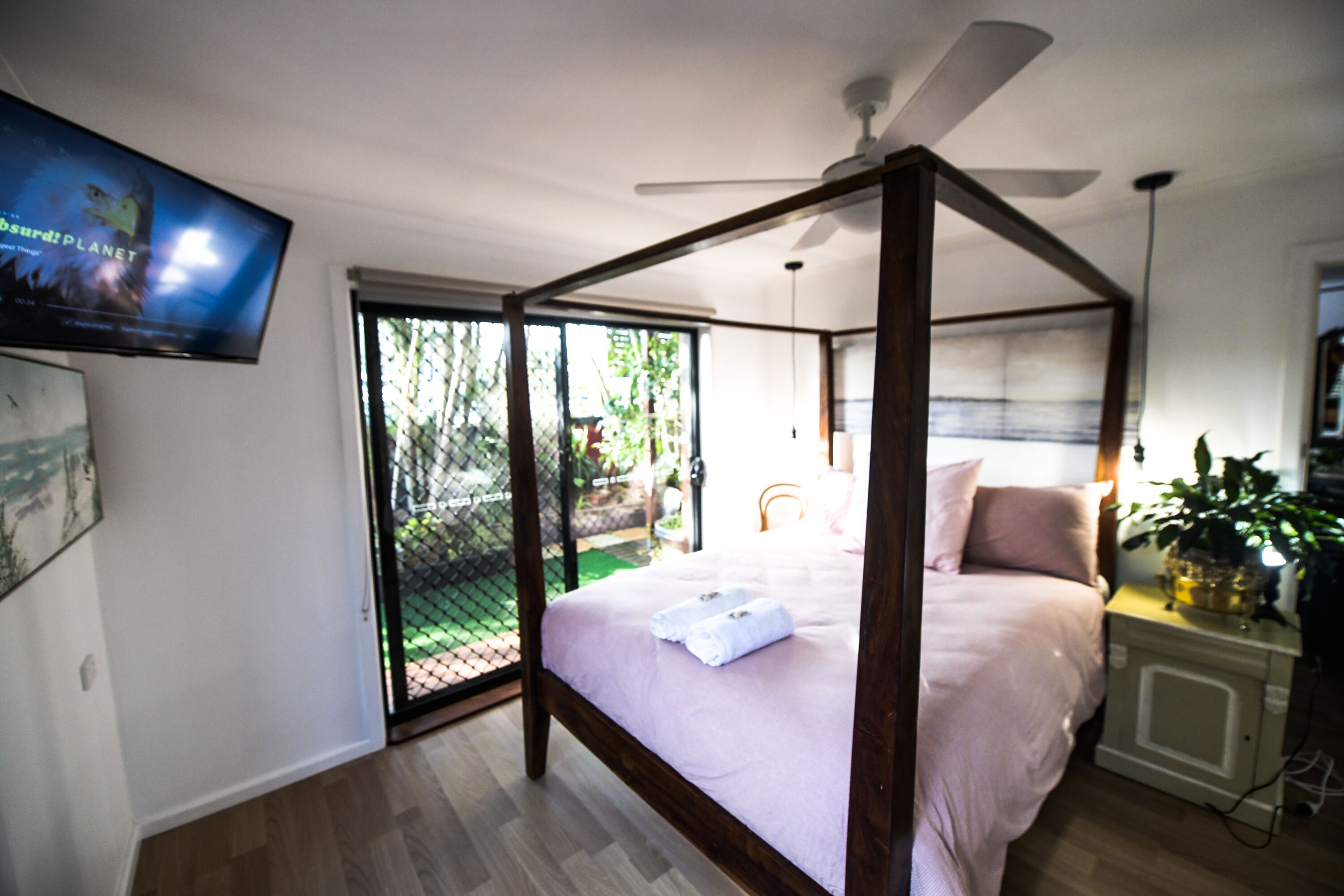 A bedroom with a four-poster bed, a ceiling fan, a TV mounted on the wall, and a sliding glass door leading to an outdoor area.