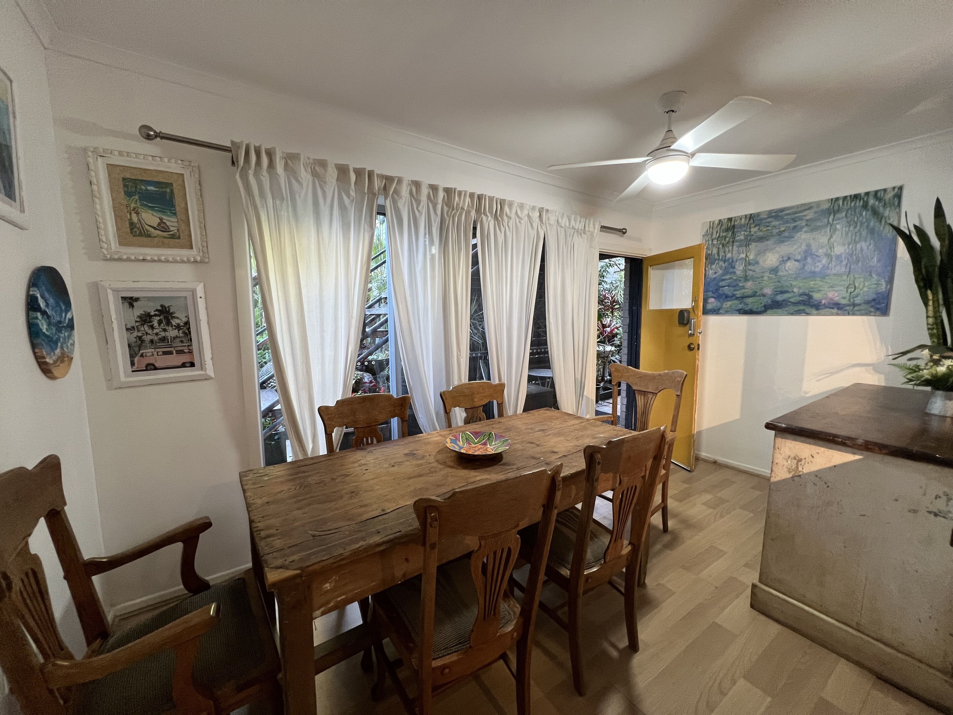 A dining area with a wooden table and chairs, a ceiling fan, and a view through curtains to an outdoor area.