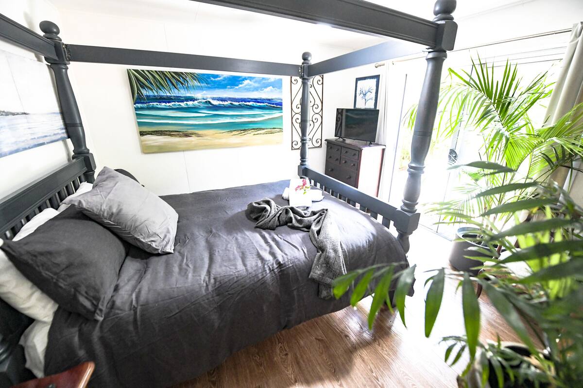 Ein Bett mit Baldachin, grau bezogen, ein Gemälde mit Strandmotiv und ein Fernseher auf einer Kommode.