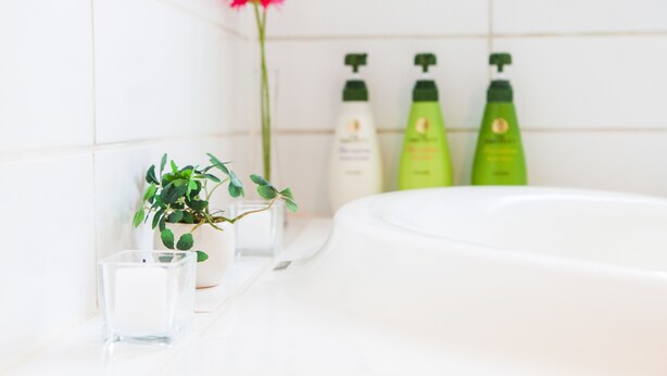 Un baño con una bañera blanca, una planta en maceta, una vela de cristal y botellas verdes de champú y acondicionador.