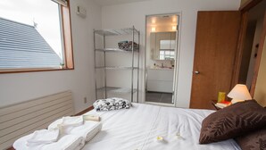Une chambre d’hôtel avec un lit, une fenêtre, une étagère en métal, une porte en bois et une table de chevet avec une lampe.