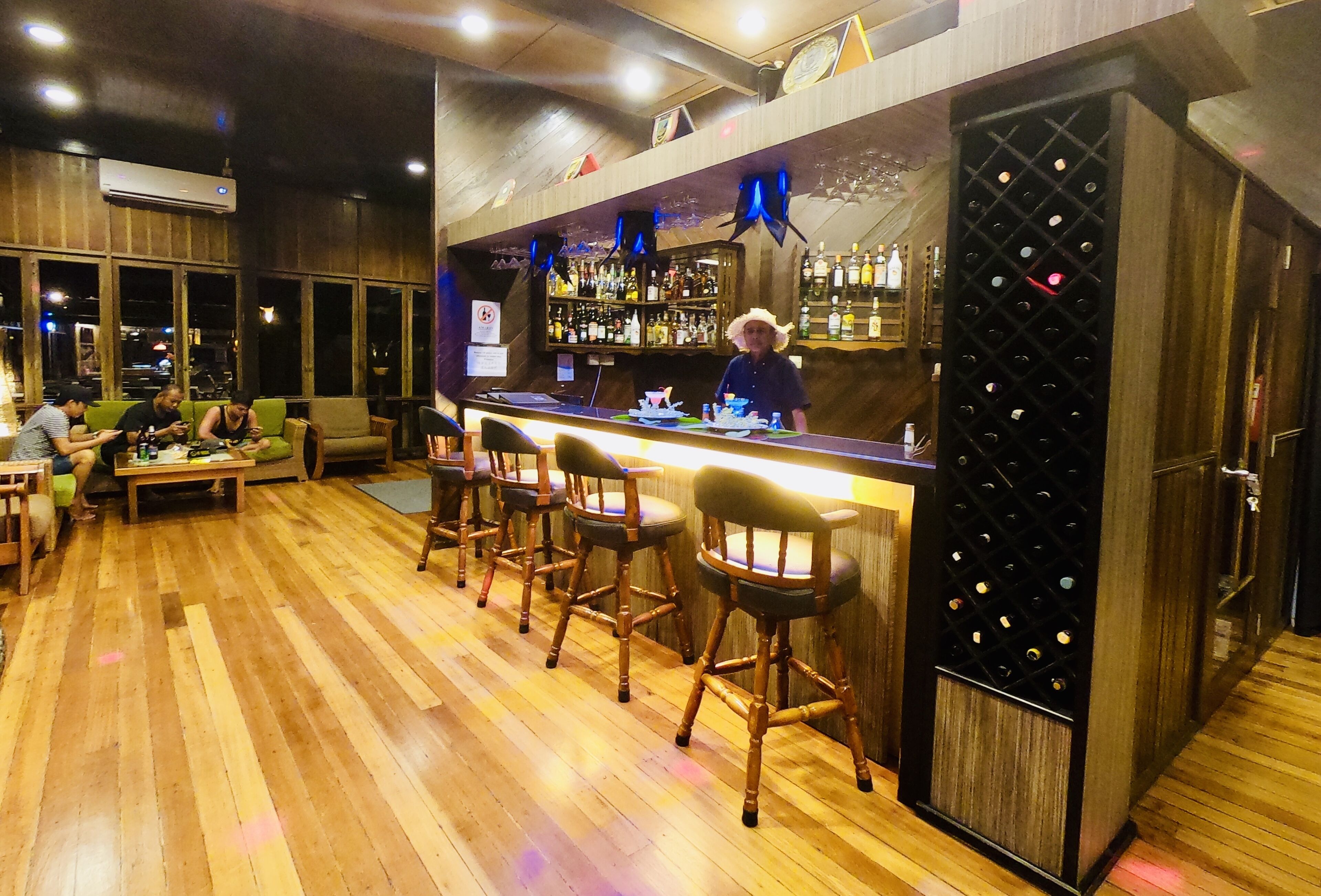 A bar area with wooden flooring, bar stools, a counter with a wine rack, and a person standing behind the bar.