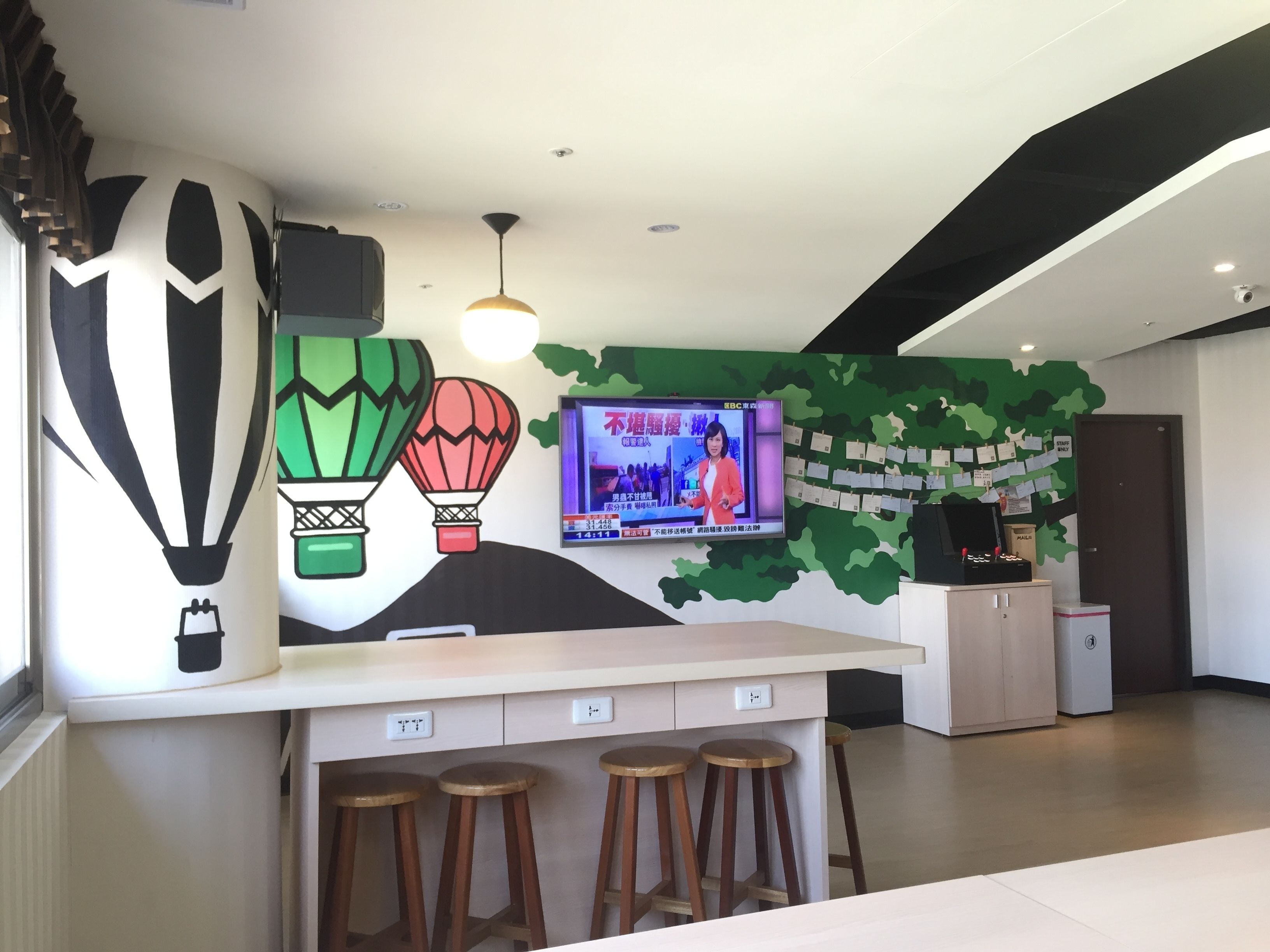 Een modern receptiegebied met een groot televisiescherm waarop een nieuwsuitzending draait, een muur met een ontwerp van groene bladeren en een bar met krukjes.