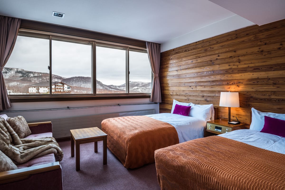 A hotel room with two beds, a wooden headboard, a small table, and a view of a mountain landscape.