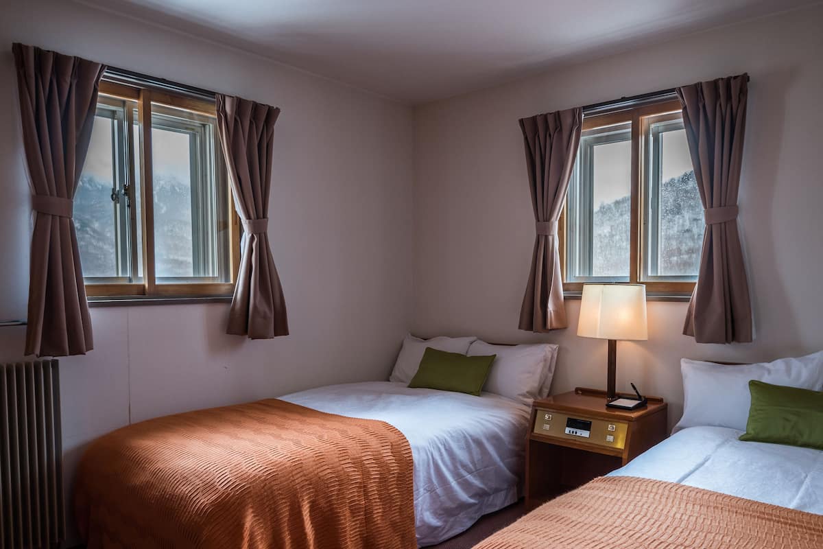 A hotel room with two single beds, a bedside table with a lamp, a window with curtains, and a radiator.