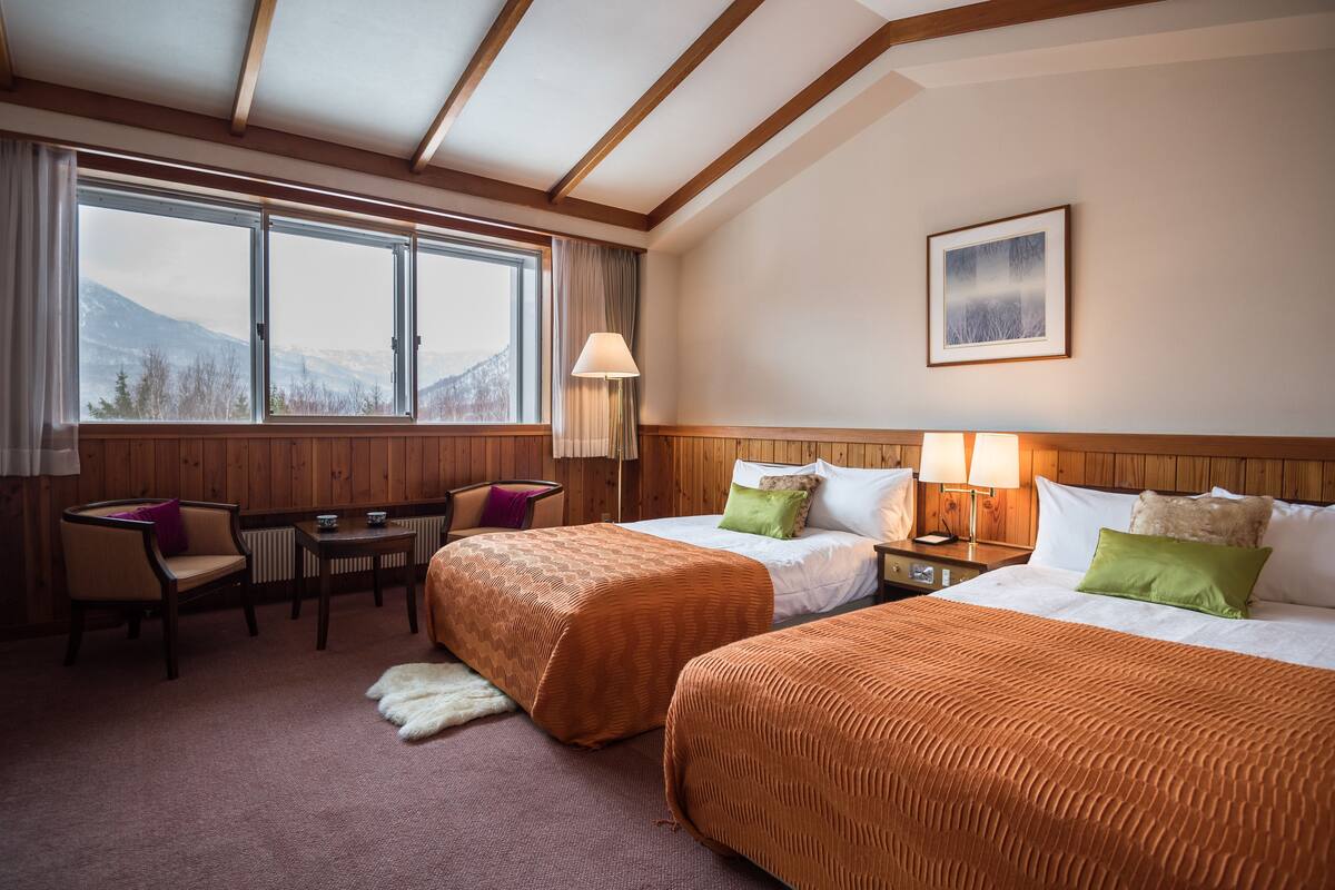 A hotel room with two beds, a desk, a chair, and a view of mountains through large windows.