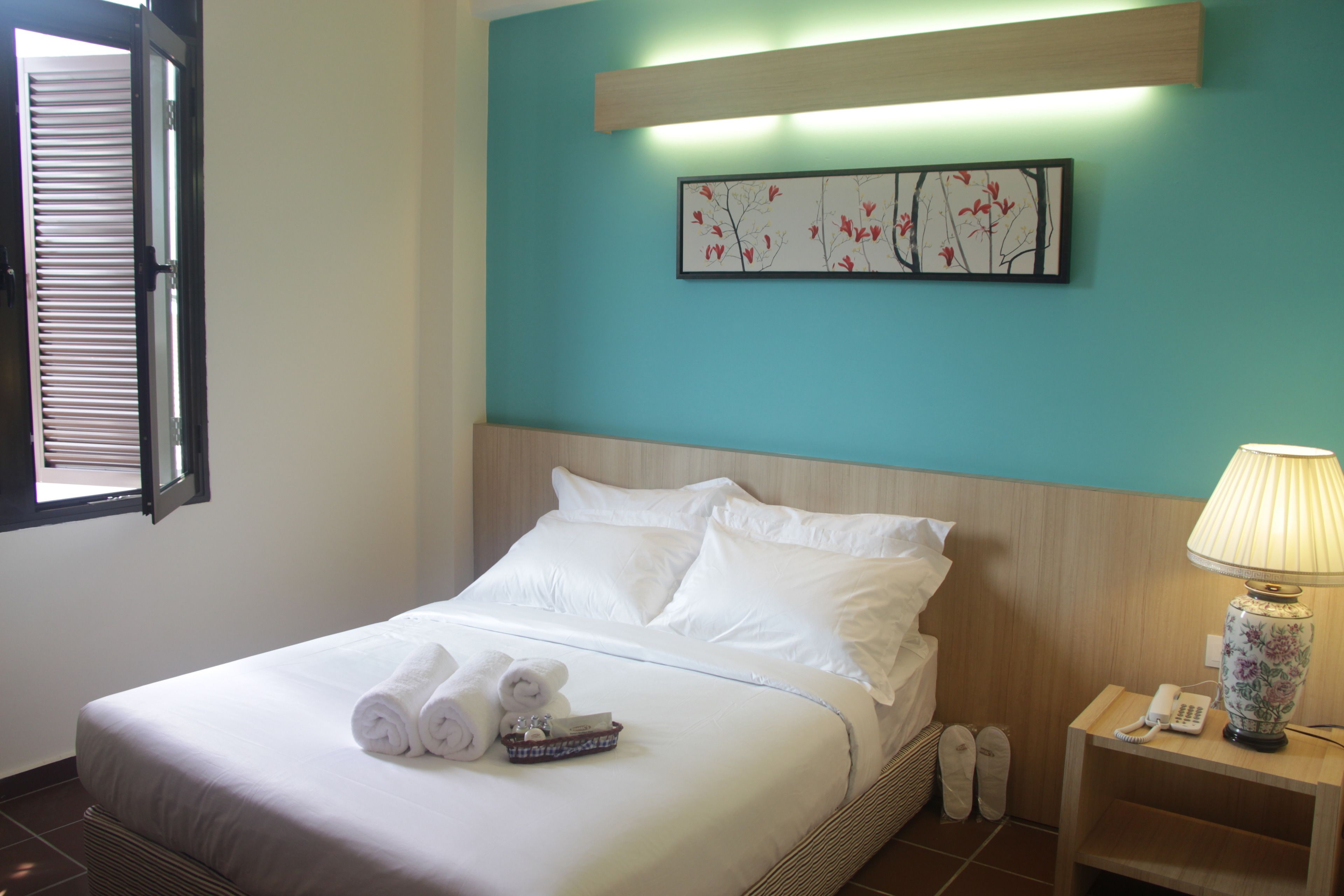 Habitación de hotel con cama, mesita de noche con lámpara, un cuadro enmarcado en la pared y una ventana con persianas.