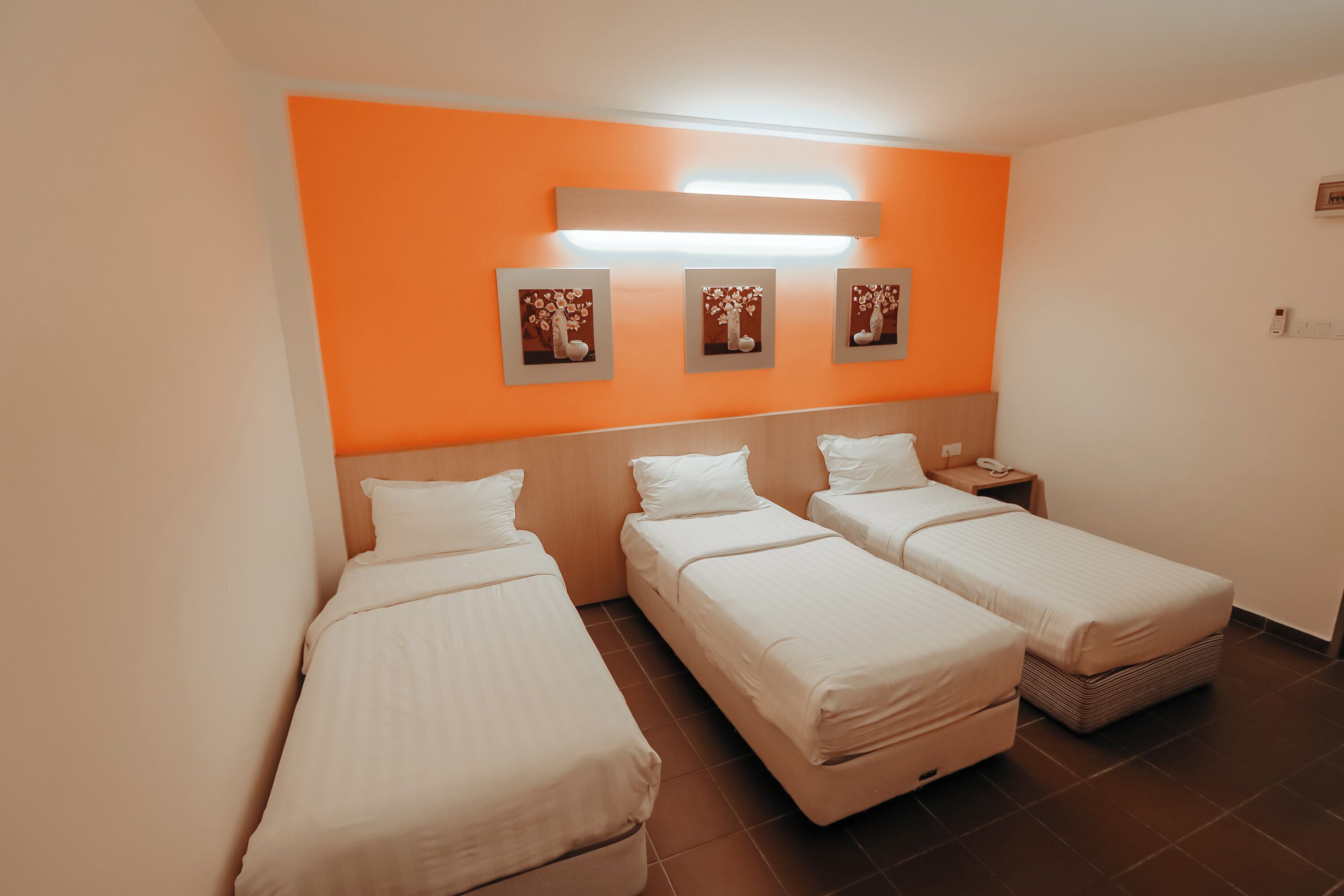 Habitación de hotel con tres camas individuales, cabecera de madera, pared de acento naranja y tres cuadros enmarcados.