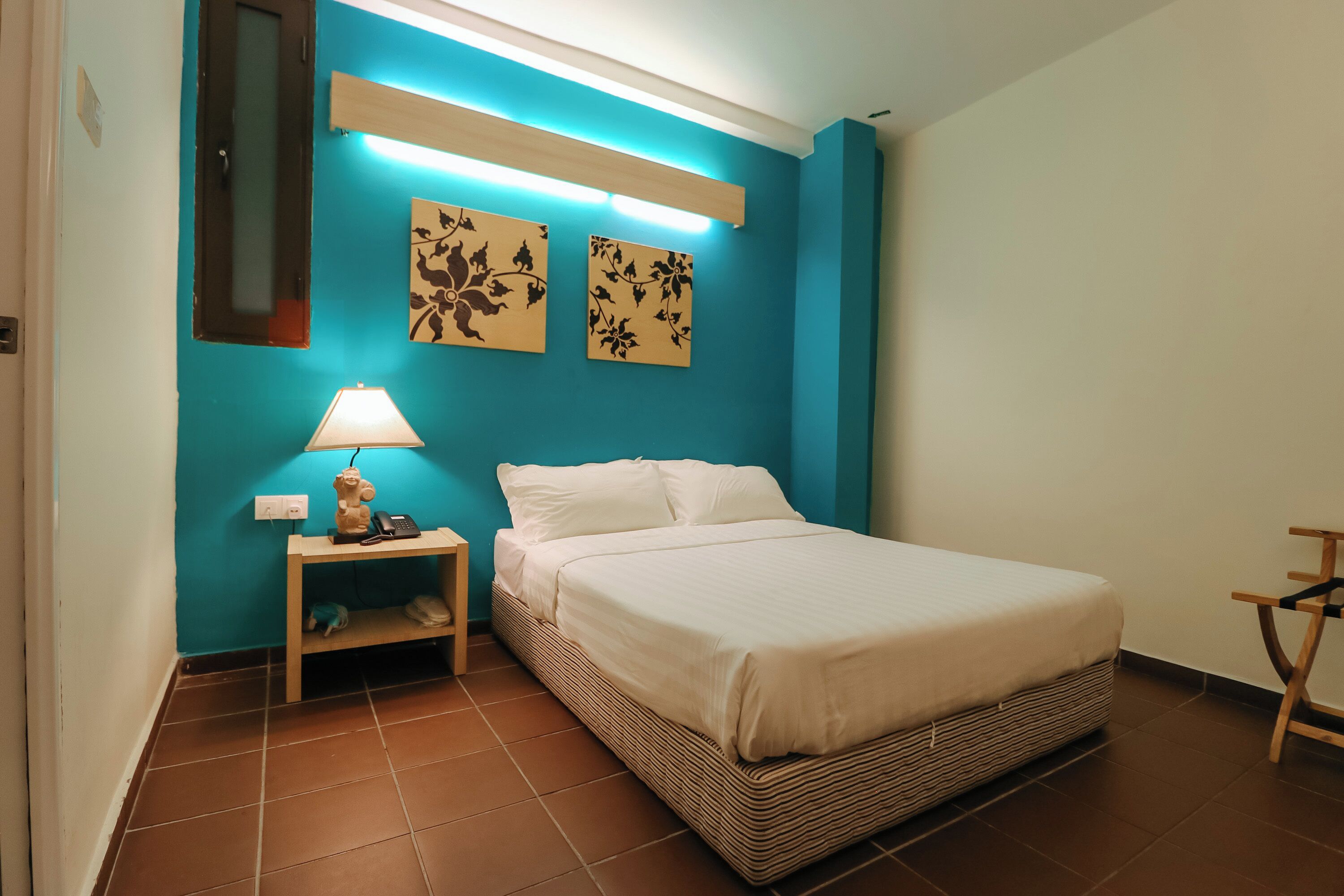 A bedroom with a bed, a nightstand with a lamp, a wall-mounted shelf, and two floral wall art pieces.