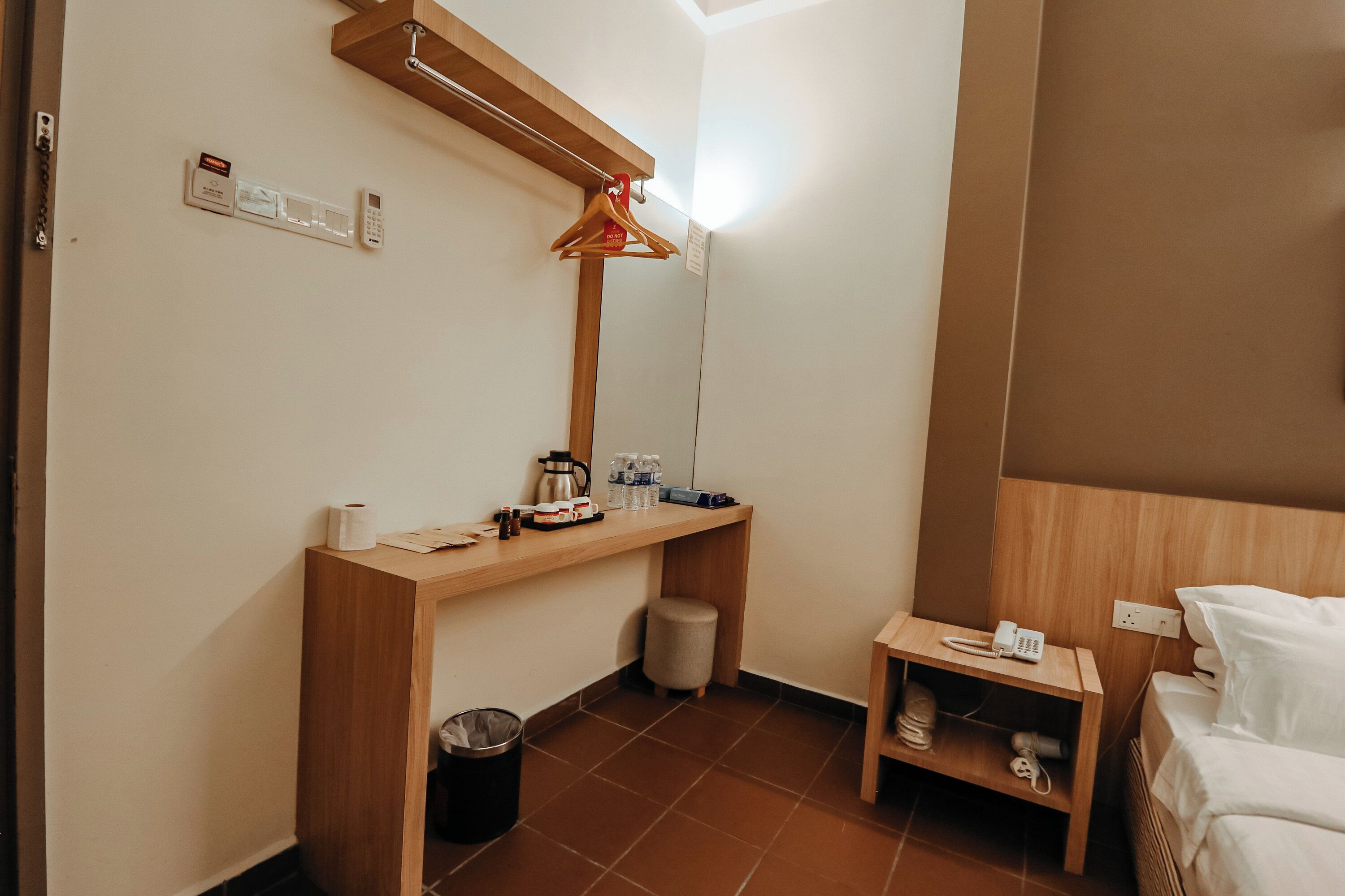 A hotel room with a wooden desk, a hanging coat rack, a small bedside table with a phone, a trash bin, and a bed with white linens.