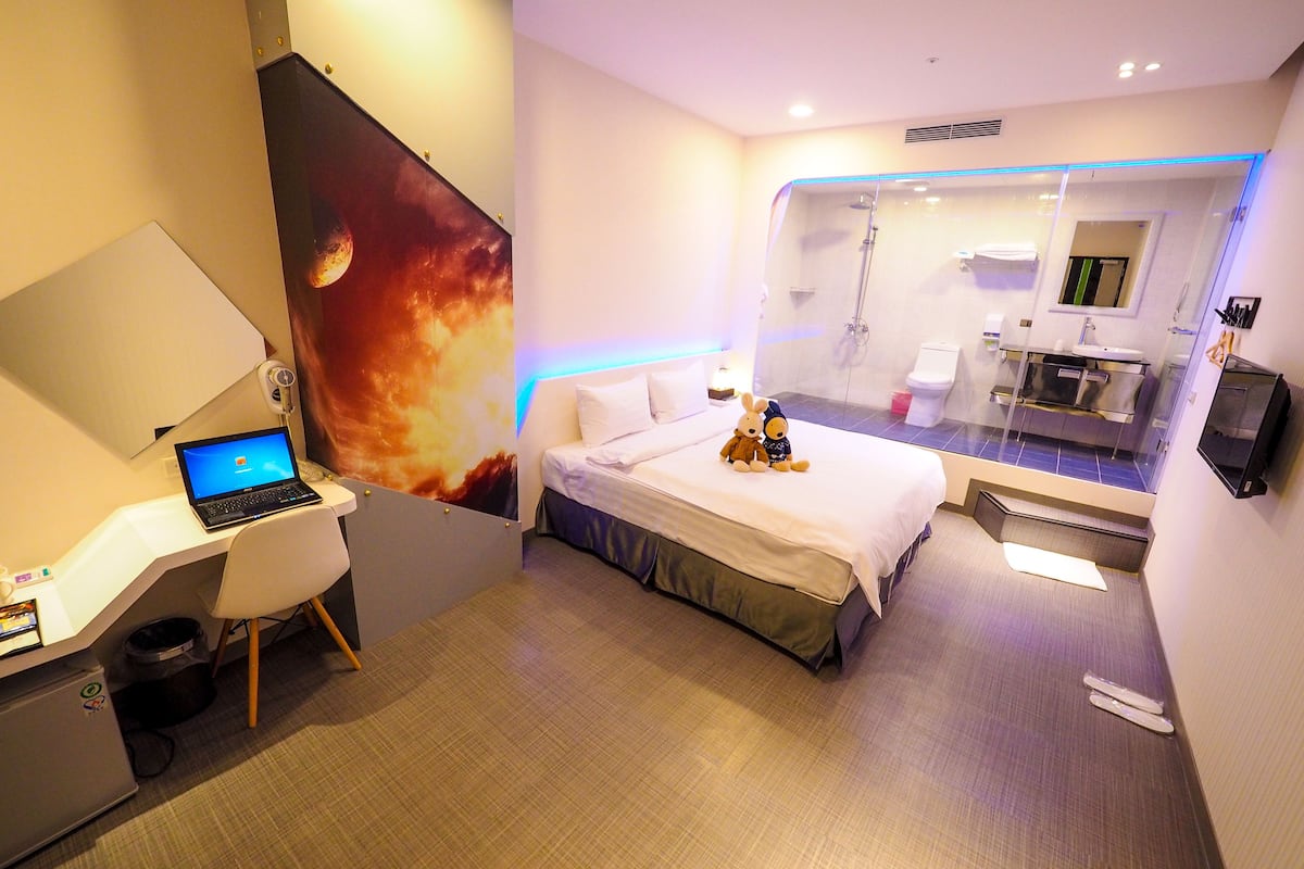 A hotel room with a bed, a desk with a laptop, a shower area, and a bathroom with a mirror.