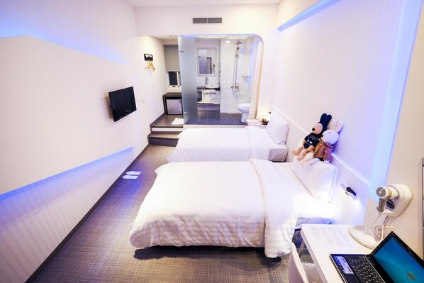 Habitación de hotel con dos camas, un escritorio con una computadora portátil, un televisor montado en la pared y un área de ducha visible a través de una puerta de vidrio.