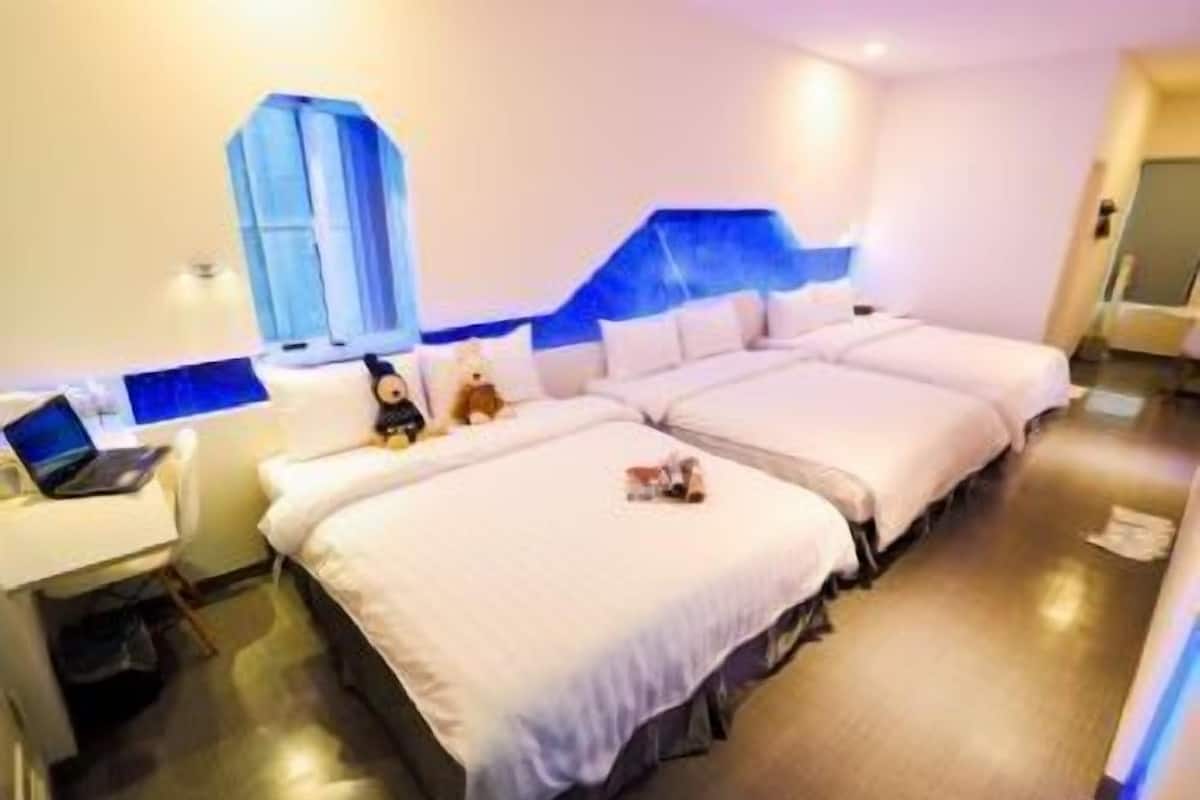 A hotel room with two beds, a desk with a laptop, and a window with a blue accent wall.