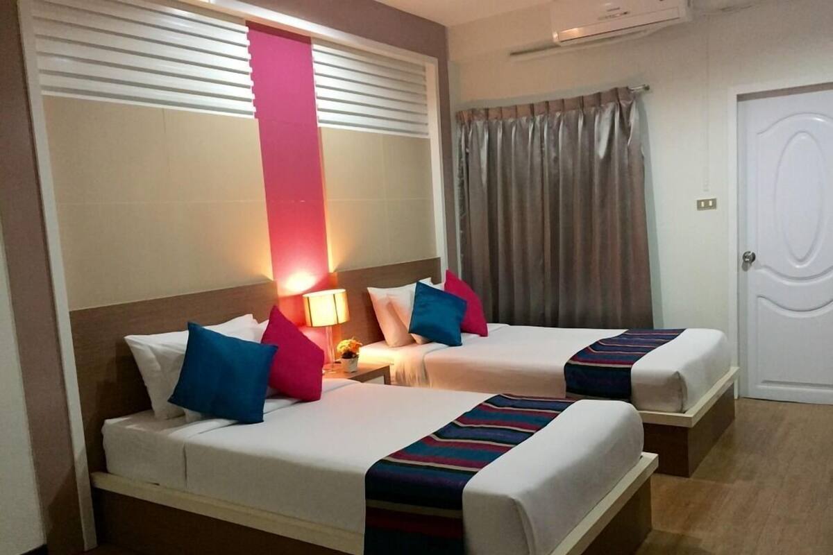 Ein Hotelzimmer mit zwei Betten, jeweils mit weißer Bettwäsche und bunten Kissen. Ein Nachttisch mit Lampe und ein kleiner Tisch mit Blumenvase sind ebenfalls vorhanden. Das Zimmer verfügt über ein großes Fenster mit Jalousien, eine Tür mit Vorhang und eine wandmontierte Klimaanlage.
