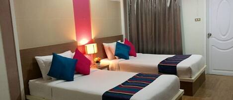 A hotel room with two beds, each with white bedding and colorful pillows. There is a nightstand with a lamp and a small table with a flower vase. The room has a large window with blinds, a door with a curtain, and a wall-mounted air conditioning unit.