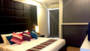 Quarto de hotel com uma cama grande, cabeceira escura, mesa de cabeceira com abajur e um aparelho de ar condicionado.