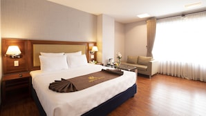 Une chambre d’hôtel avec un grand lit, une table de chevet avec une lampe, une petite table avec un vase de fleurs, un canapé et une fenêtre avec des rideaux.