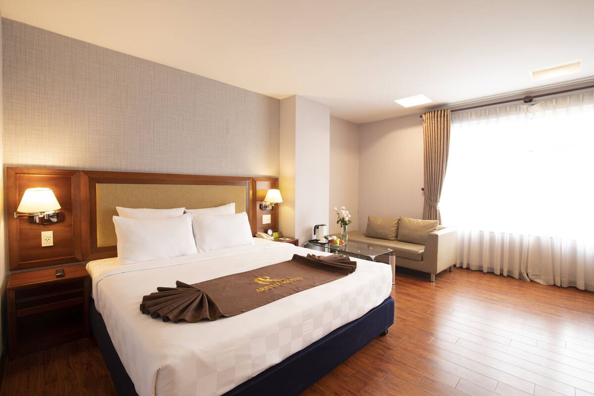 Une chambre d’hôtel avec un grand lit, une table de chevet avec une lampe, une petite table avec un vase de fleurs, un canapé et une fenêtre avec des rideaux.