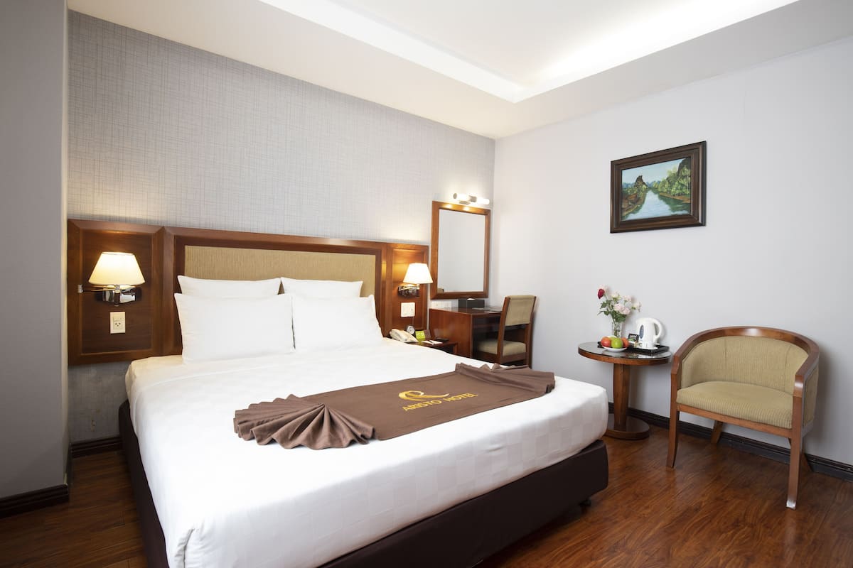 Une chambre d’hôtel avec un grand lit, une tête de lit en bois, une table de chevet avec une lampe, une chaise, une petite table avec un vase de fleurs et un tableau encadré au mur.