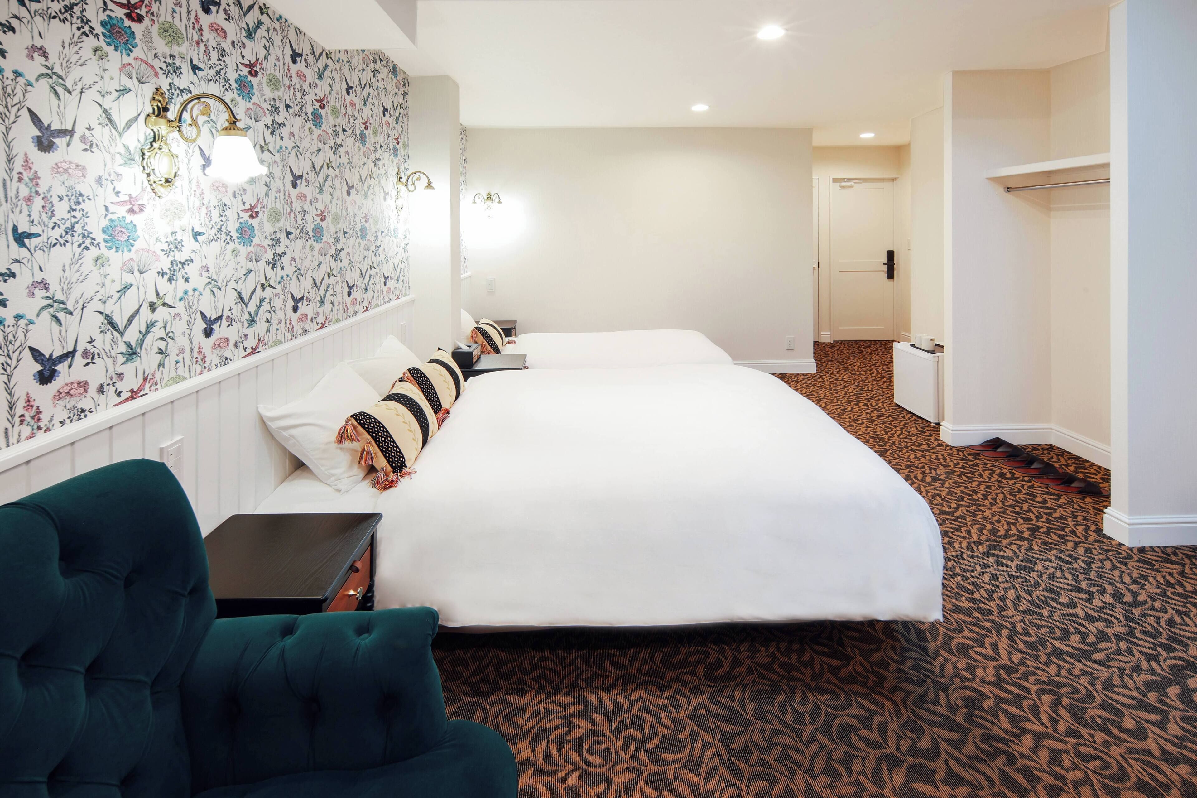A hotel room with two beds, a teal armchair, a small table, and a patterned carpet.