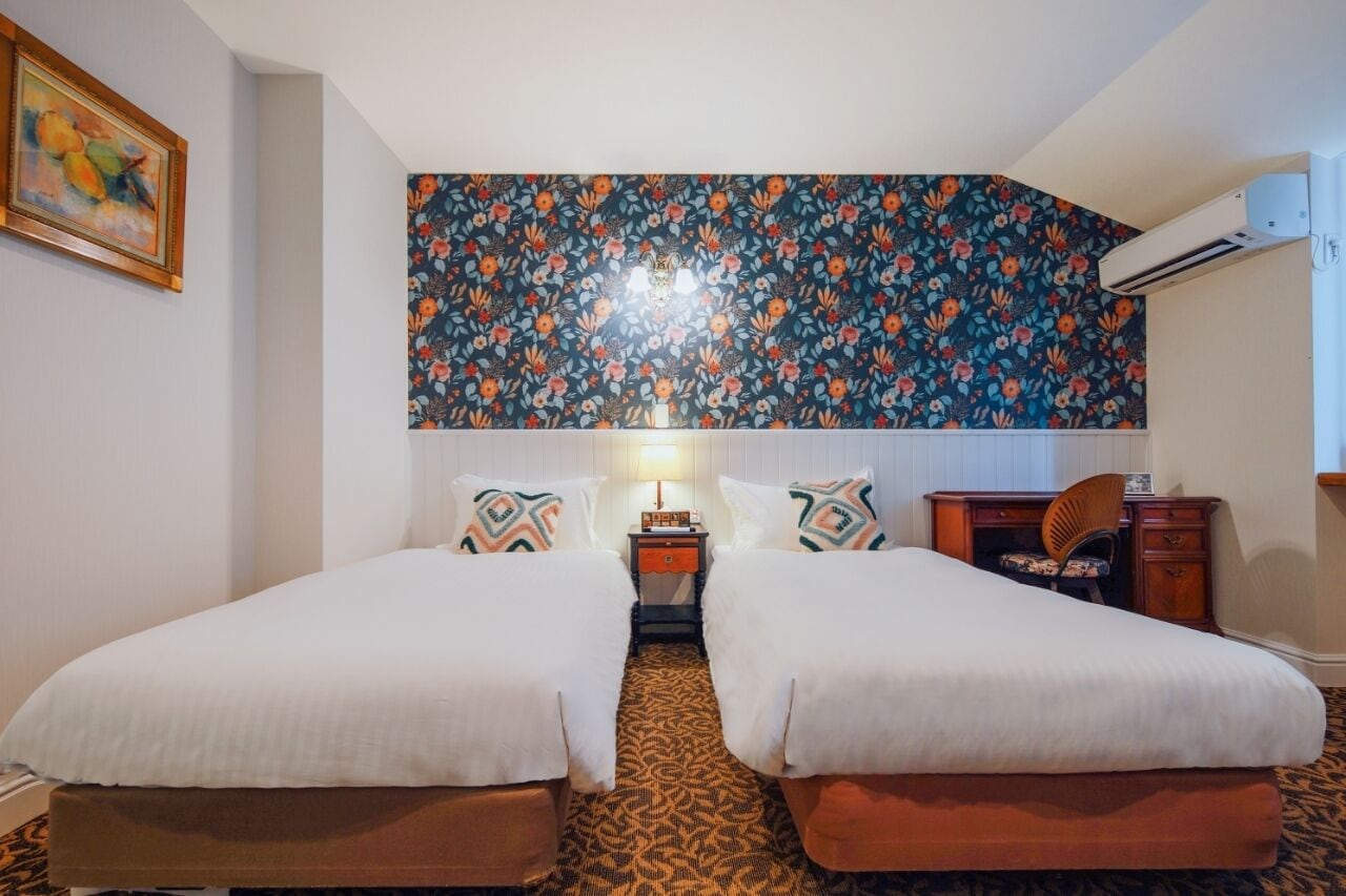 A hotel room with two beds, a desk, a chair, a nightstand, and a floral-patterned wall.