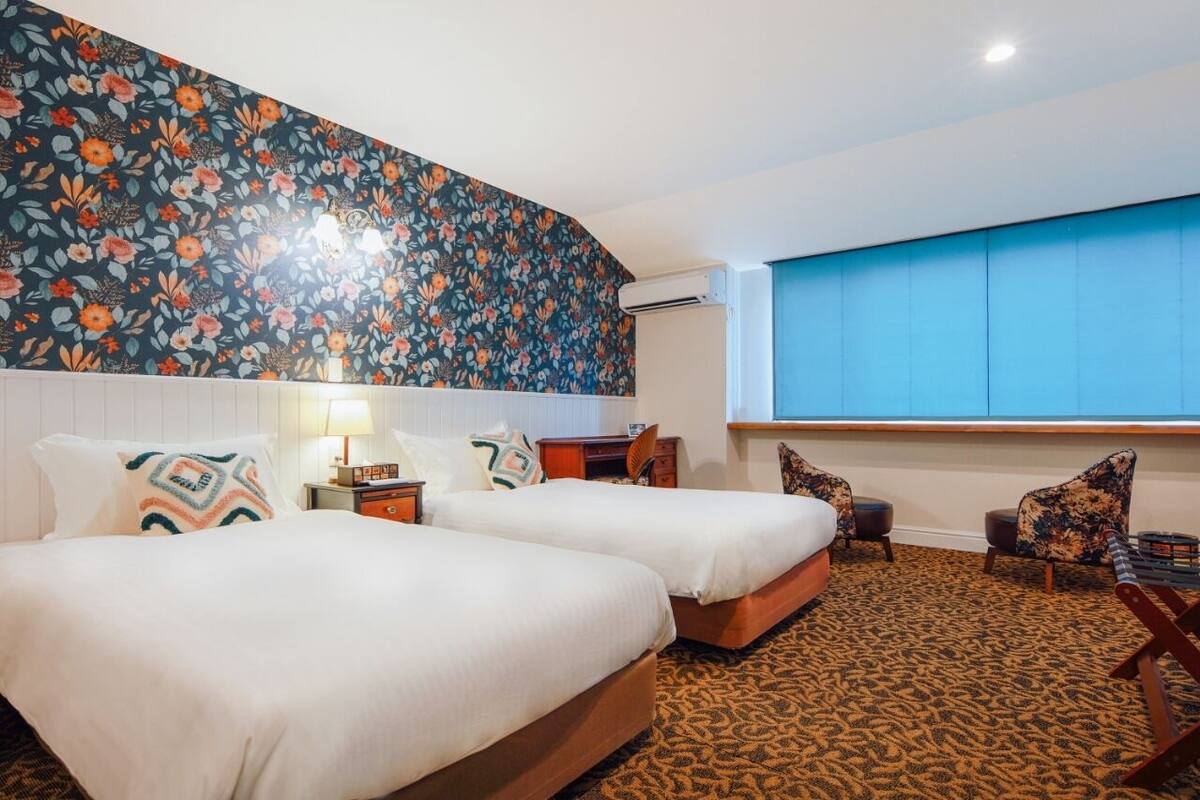 A hotel room with two beds, a desk, a chair, and a large window with blue blinds.
