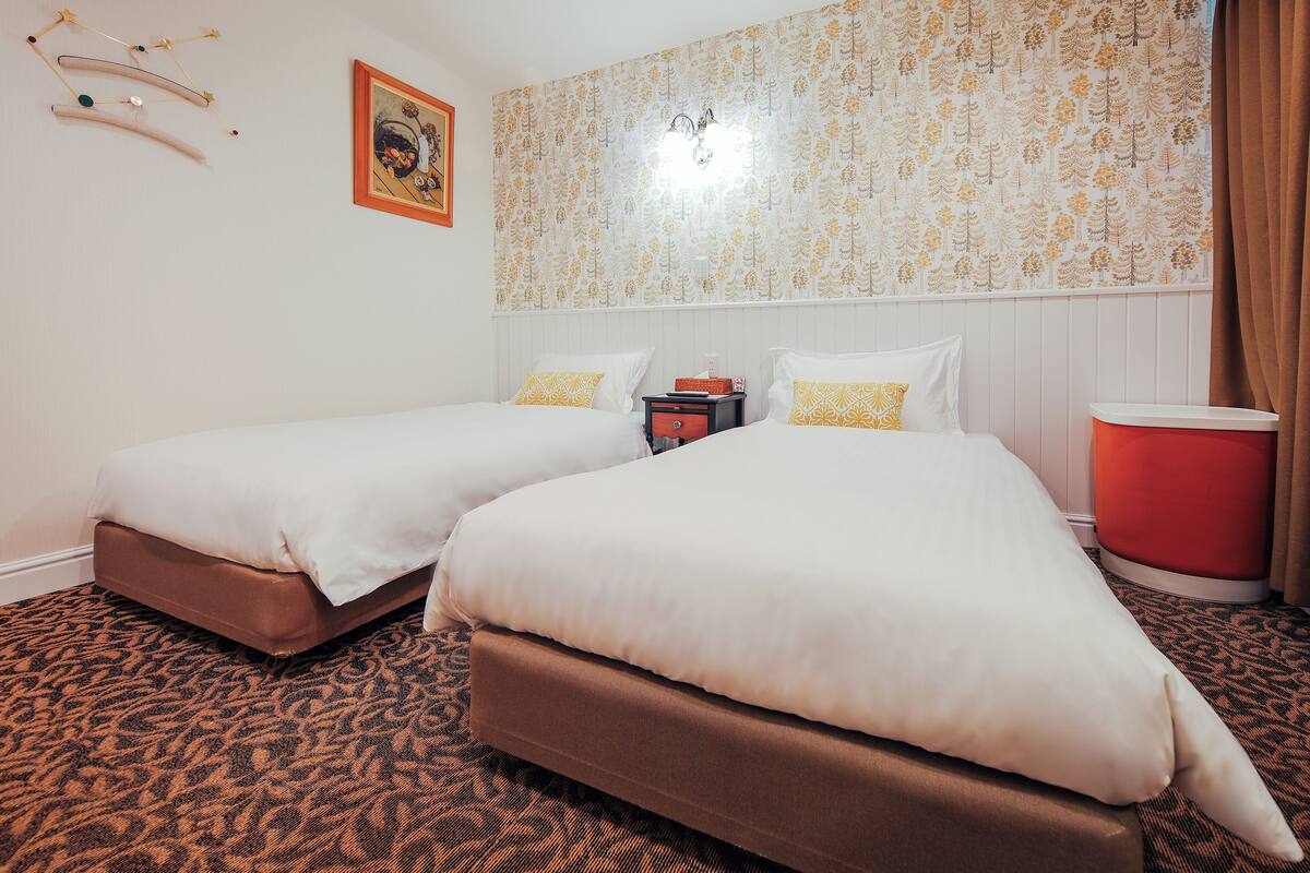 A hotel room with two beds, a patterned carpet, a red trash bin, and a wall with a floral wallpaper.