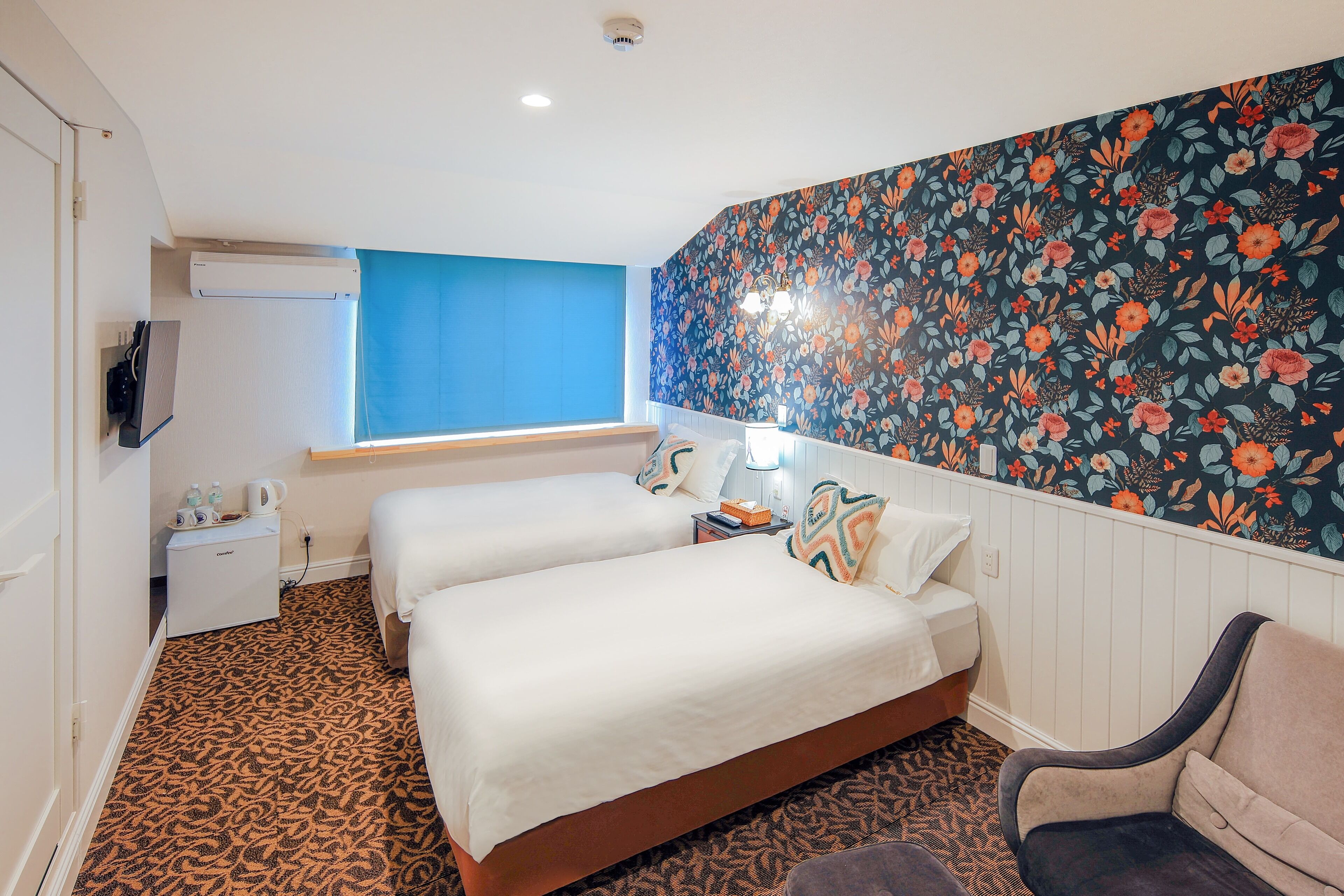 A hotel room with two beds, a desk with a lamp, a chair, a window with blue curtains, and a floral wallpaper.