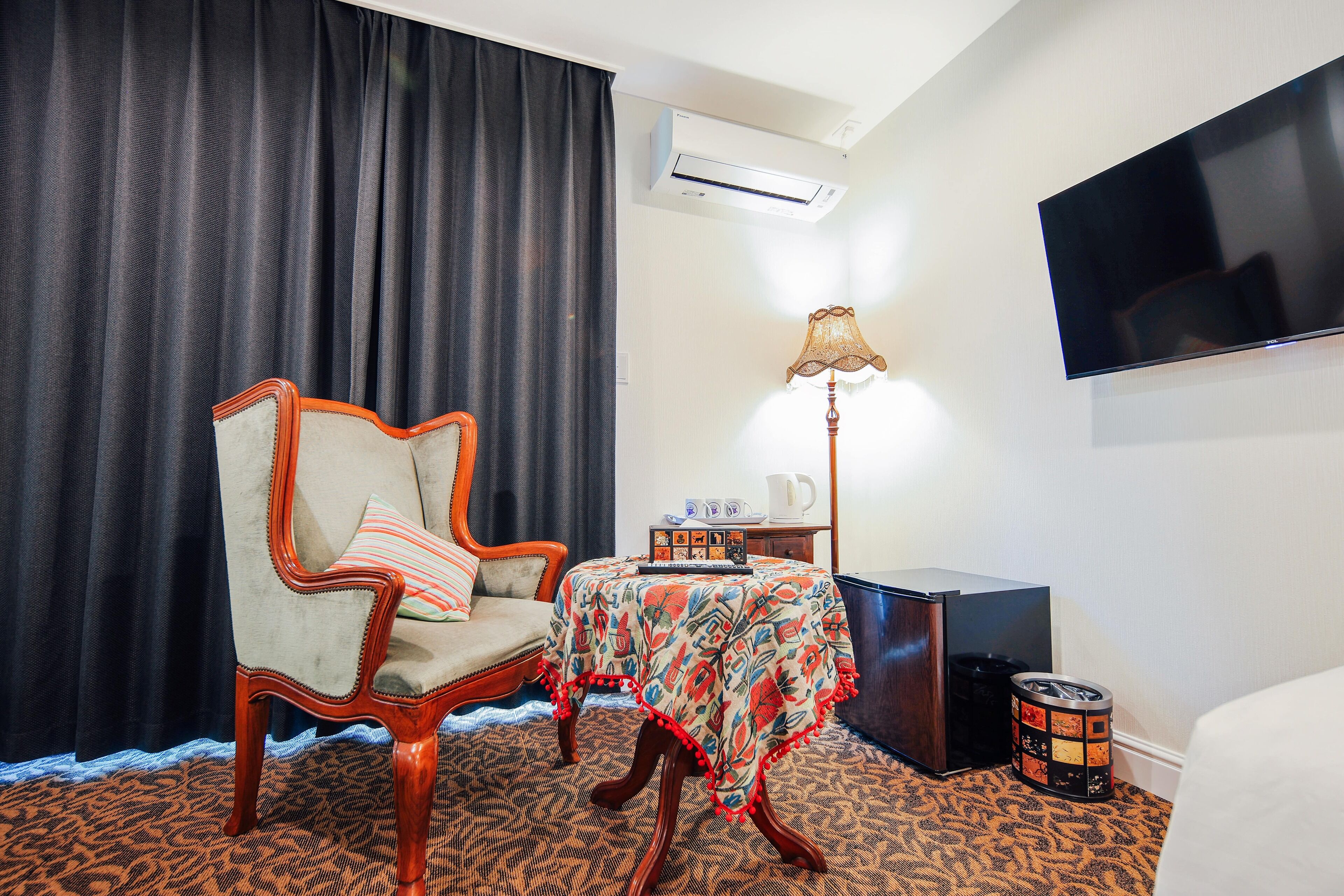 A hotel room with a large bed, a chair, a small table with a lamp, a TV, and an air conditioning unit.