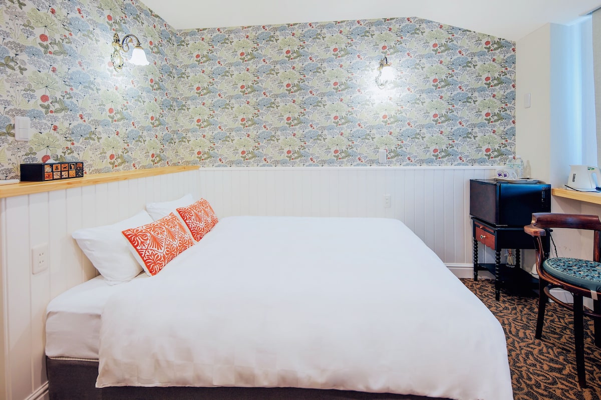 A neatly made bed with white and orange pillows, a wooden nightstand with a clock, a small table with a lamp, and a chair with a patterned cushion.