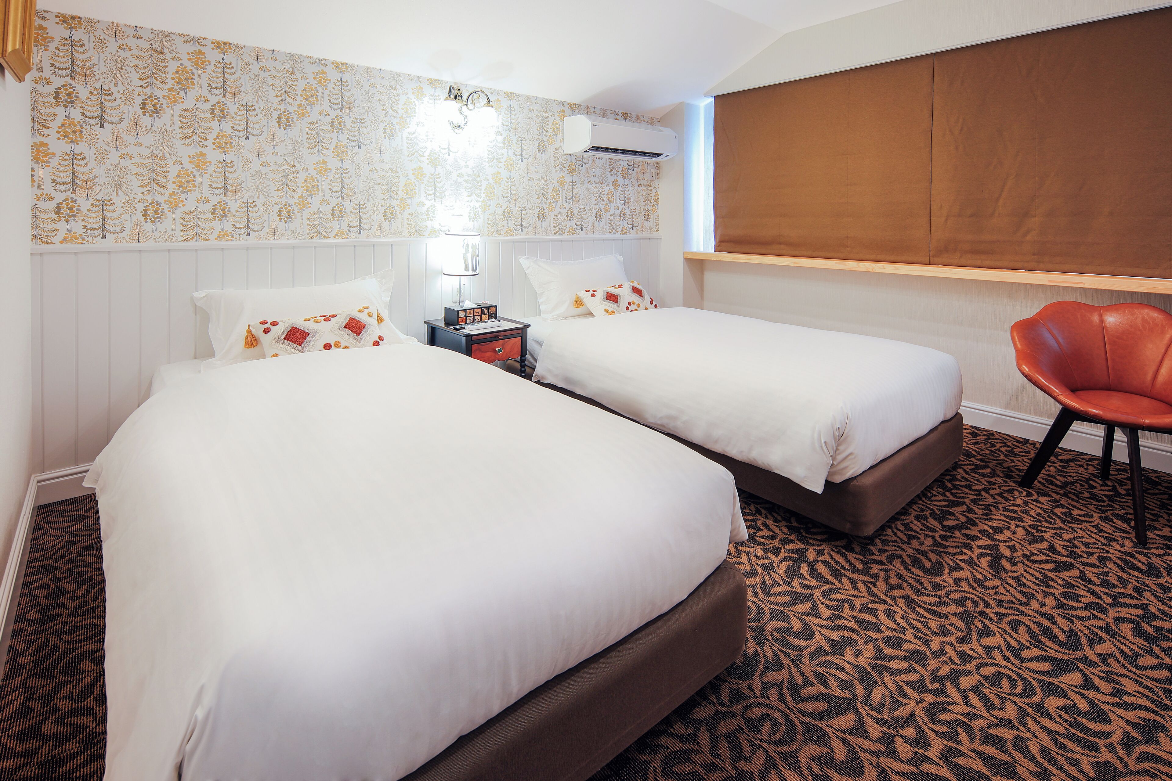 A hotel room with two beds, a patterned carpet, a wooden headboard, a red chair, and a wall-mounted air conditioner.