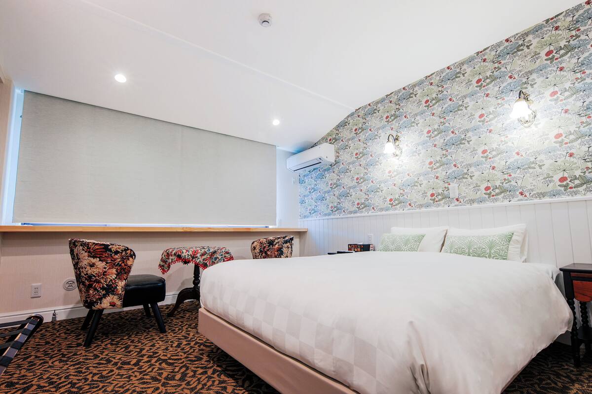 A hotel room with a large bed, a desk, and a chair. There is a window with a view of the outside.