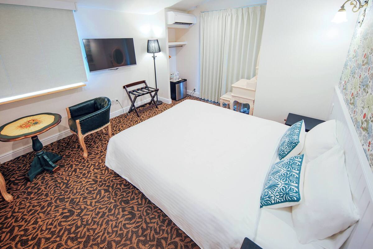 A hotel room with a large bed, a small table, a chair, a TV, and a window with curtains.