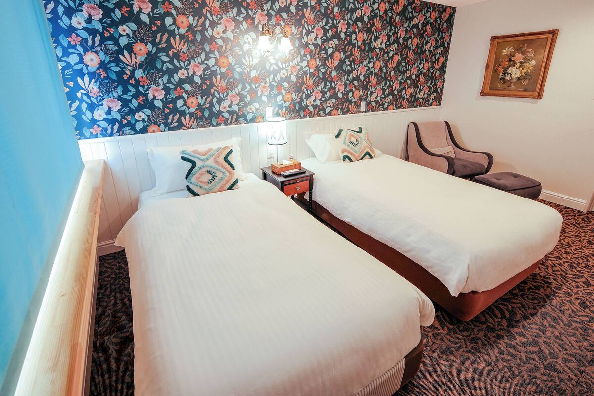 A hotel room with two beds, a nightstand, a lamp, a chair, and a floral wallpaper.