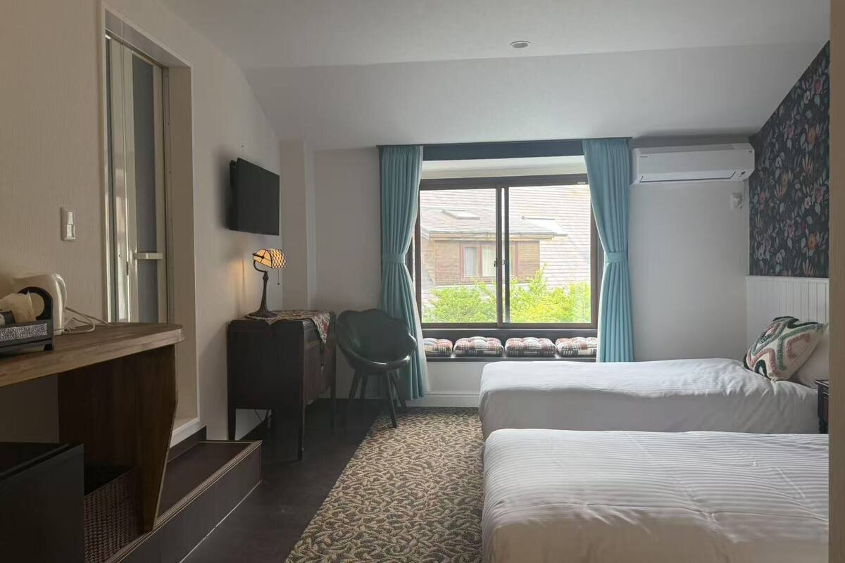 A hotel room with two beds, a desk with a lamp, a chair, and a large window with curtains.