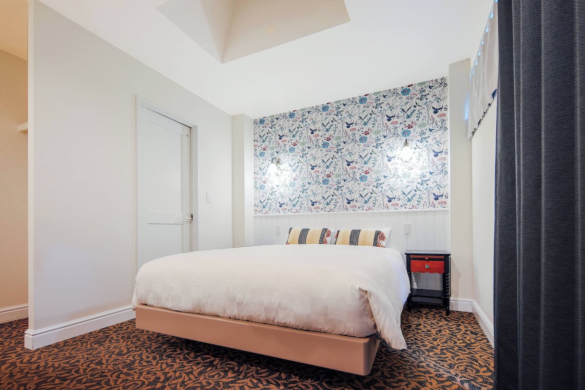 A bedroom with a bed, a patterned wall, a door, and a small table with a red drawer.