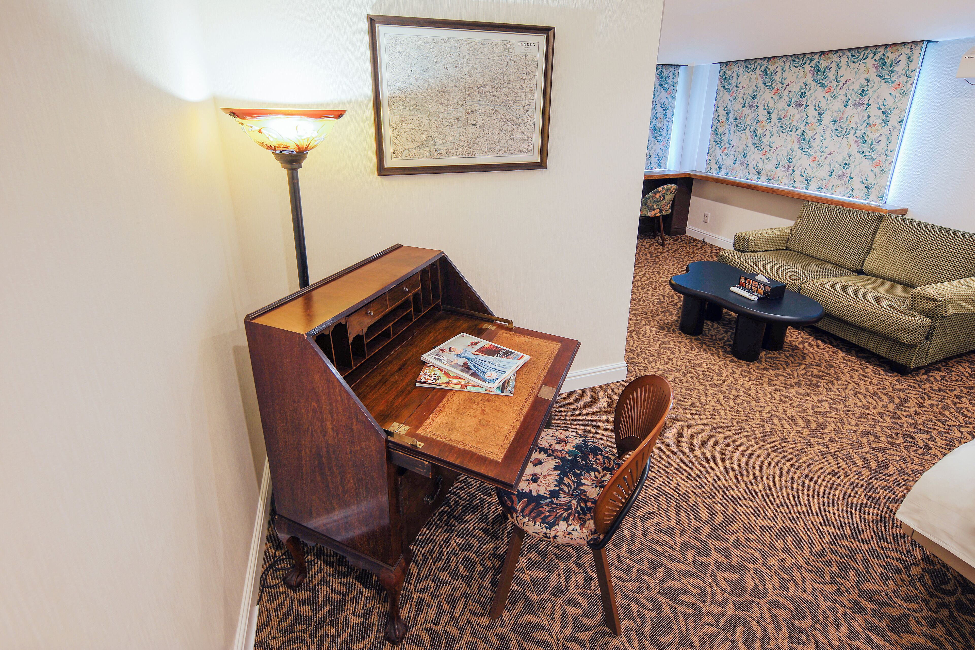 A hotel room with a wooden desk, a lamp, a map on the wall, a patterned carpet, a sofa, and a window with floral wallpaper.