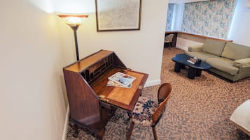A hotel room with a wooden desk, a lamp, a map on the wall, a patterned carpet, a sofa, and a window with floral wallpaper.