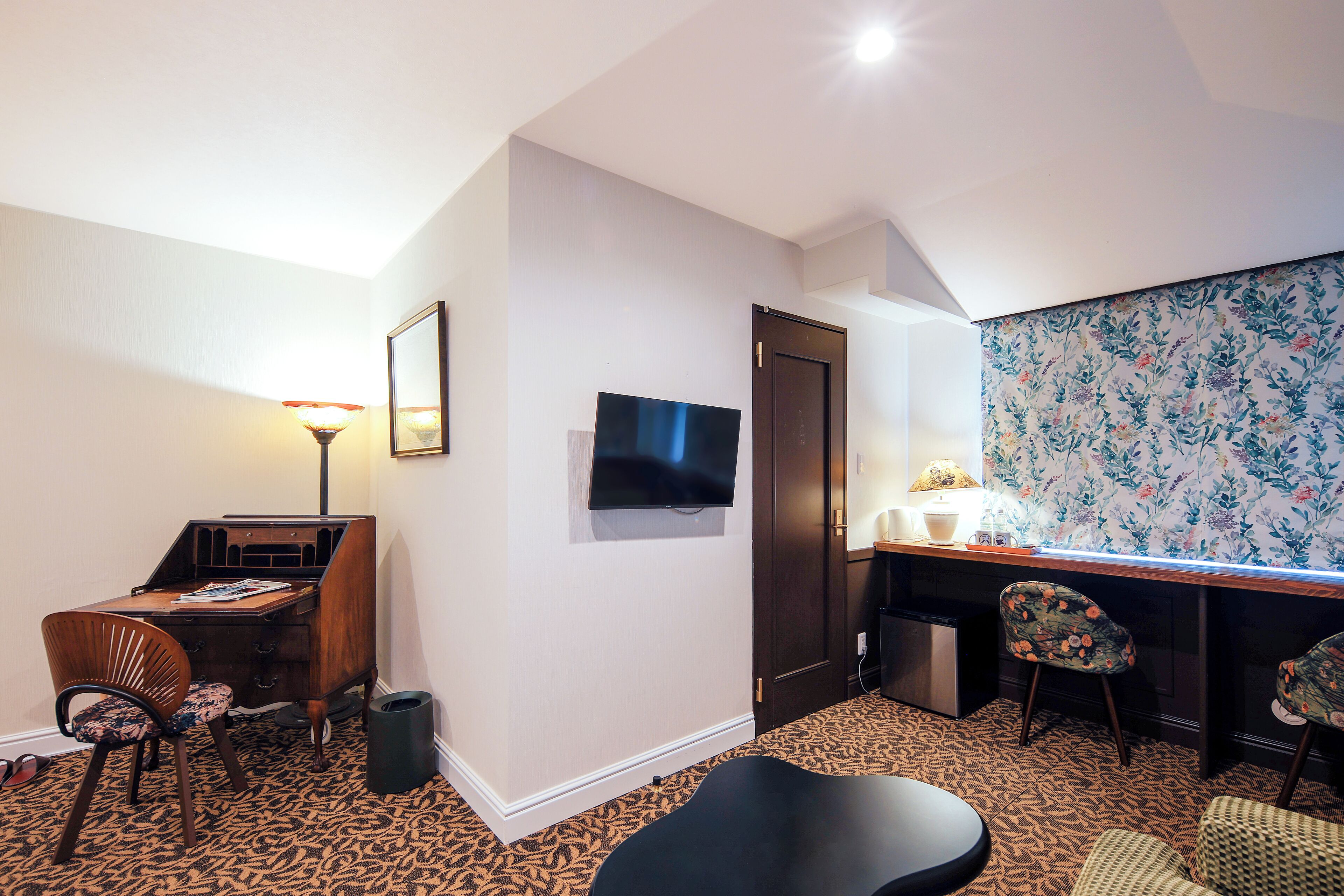 A compact hotel room with a desk, a television, a patterned carpet, and a small seating area.