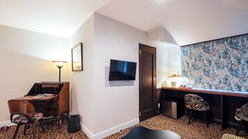A compact hotel room with a desk, a television, a patterned carpet, and a small seating area.