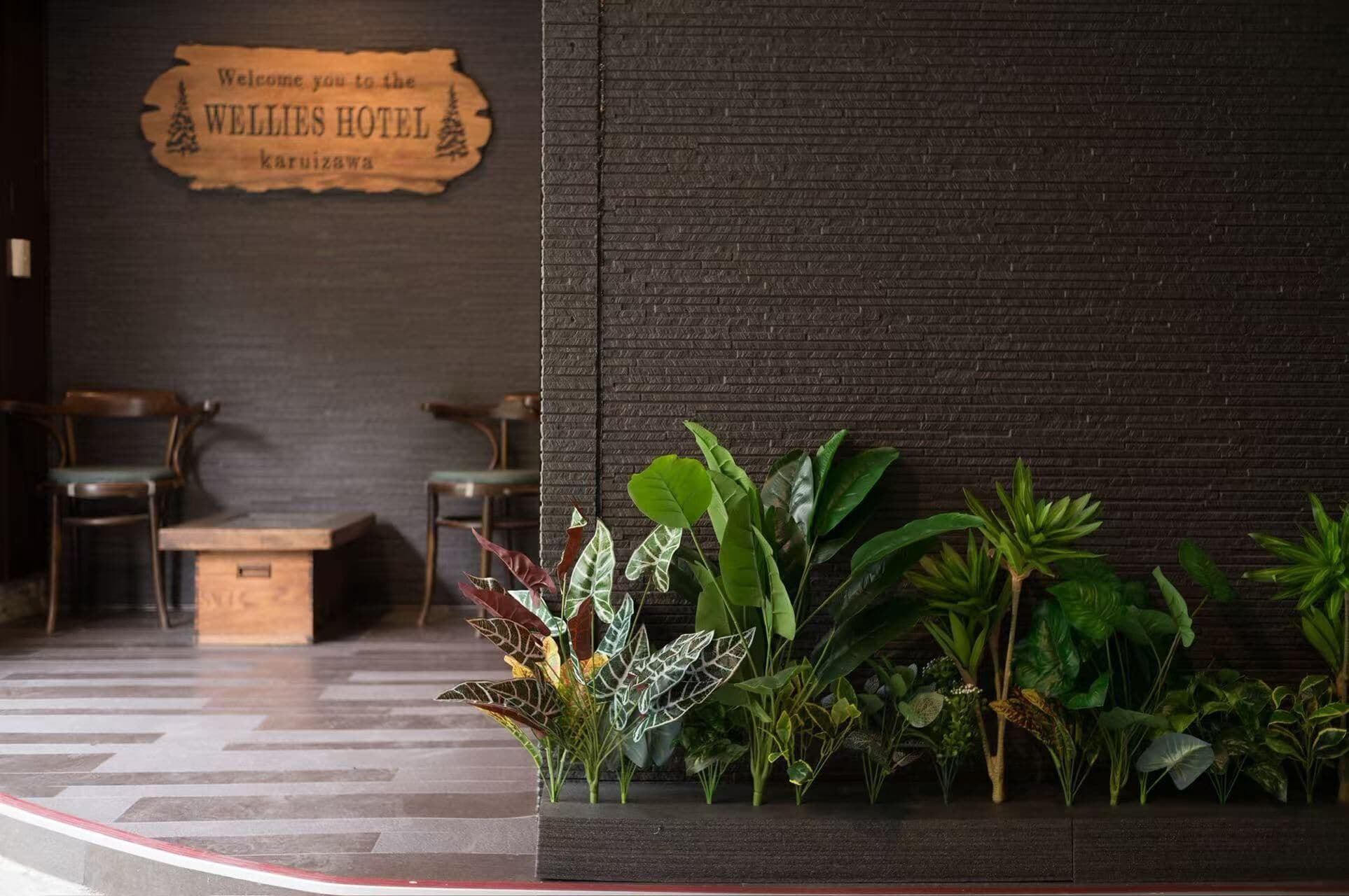 Et receptionsområde med et træskilt, der skriver 'Welcome you to the WELLIES HOTEL Karuizawa', en bænk, to stole og et udvalg af grønne planter.