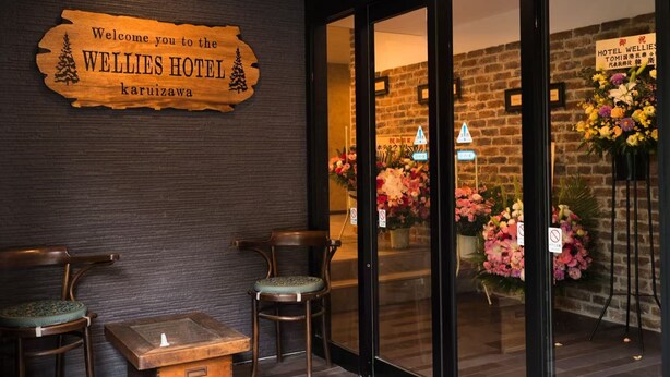 The exterior of the Wellies Hotel with a wooden signboard, two wooden chairs, and a flower arrangement outside the entrance. - Hotel WELLIES (Karuizawa)