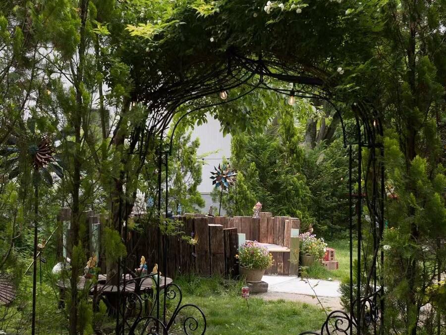 A garden with a wooden pergola, metal chairs, and a pathway leading to a building.
