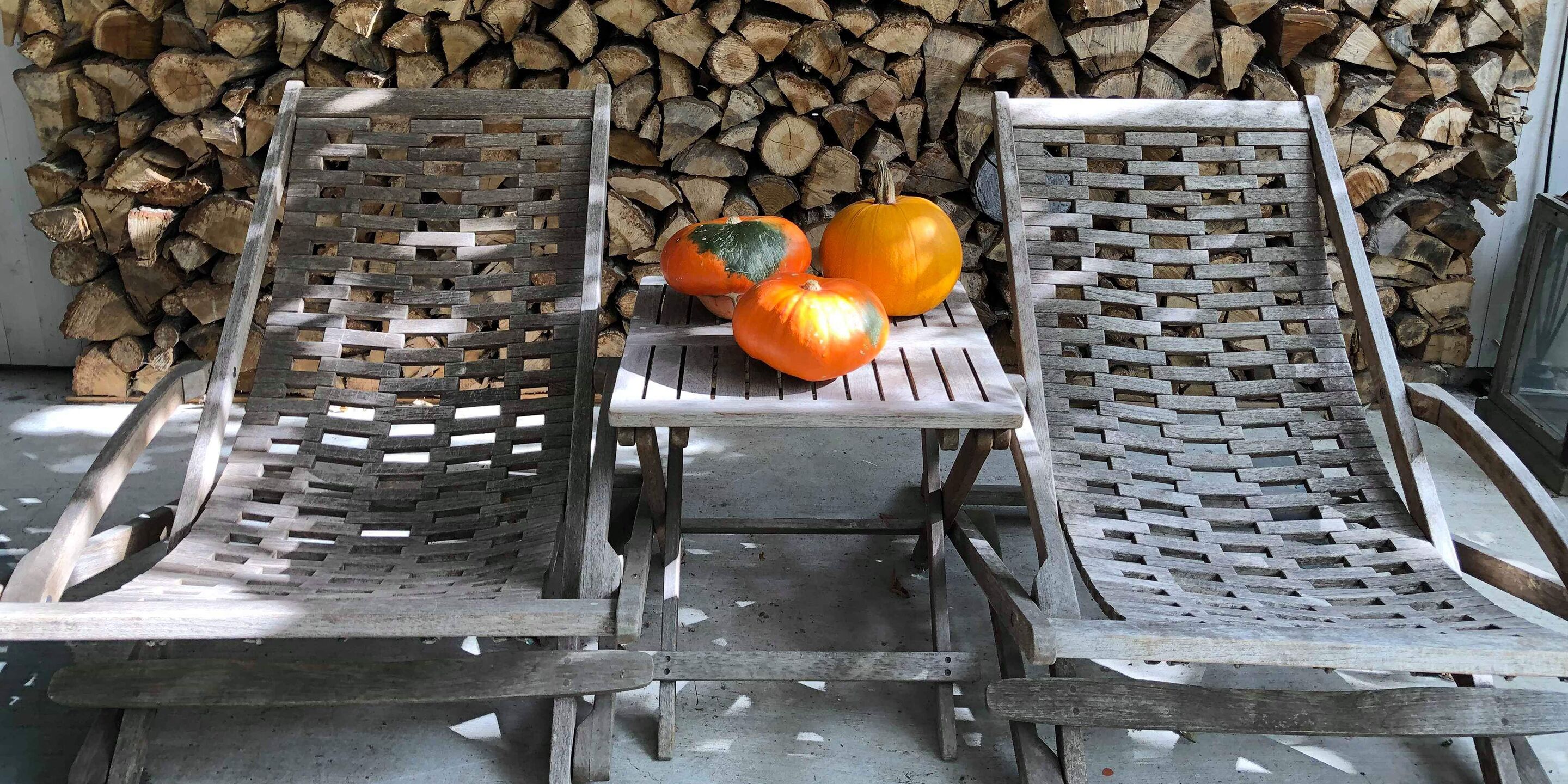 Zwei Holz-Loungesessel mit gewebtem Rücken, ein kleiner Tisch und drei Kürbisse vor einem Holzstoß.